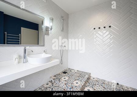 Bagno luminoso con piastrelle a spina di pesce bianche, pavimento a motivi e mobile da lavabo sospeso con lavandino ovale Foto Stock