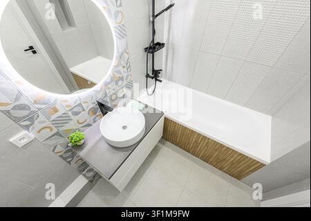 bagno moderno con specchio rotondo illuminato, lavandino sospeso, vasca da bagno e piastrellatura elegante in grigio e a fantasia Foto Stock
