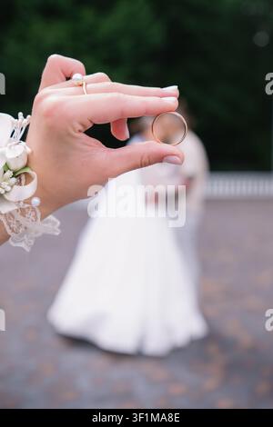 Un primo piano che si concentra sulla mano della sposa che tiene un anello nuziale d'oro. Lo sfondo mostra una silhouette romantica e sfocata della sposa e gr Foto Stock