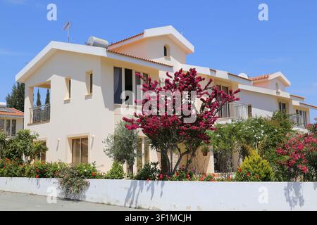 Una villa a due piani color crema con balconi è incorniciata da un bougainvillea rosa in piena fioritura e da un lussureggiante giardino a Cipro. Foto Stock