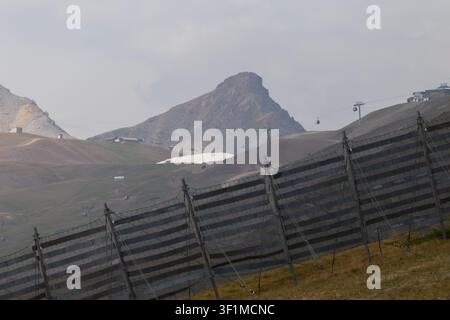 Stazione sciistica alpina in stagione estiva con recinzioni per la ritenzione della neve e linee di teleferica sotto un cielo nuvoloso Foto Stock