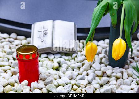 Candela commemorativa rossa e tulipani gialli su ciottoli bianchi accanto a una scultura di libro in pietra aperta su una tomba in un cimitero Foto Stock