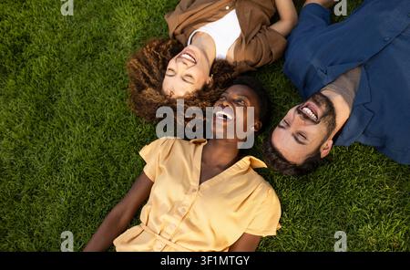 Vista dall'alto di tre amici diversi sdraiati sull'erba verde e che ridono insieme, godendosi una giornata soleggiata e rilassata all'aperto, celebrando l'amicizia, la gioia e Foto Stock
