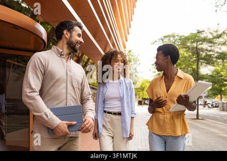 Tre colleghi camminano e parlano all'aperto accanto a un moderno edificio per uffici, tenendo un tablet, condividendo idee durante un incontro informale in una giornata di sole. Foto Stock