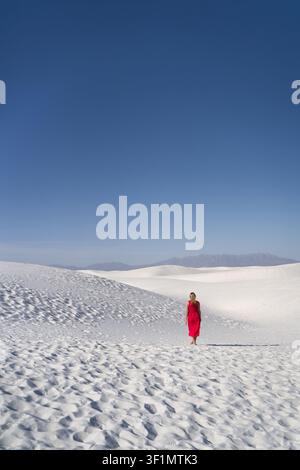 Donna in abito rosso cammina da sola su dune di sabbia bianca brillante negli USA, creando un contrasto sorprendente contro il cielo blu chiaro, che trasmette viaggio e fr Foto Stock