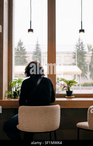 Veduta posteriore di una persona solitaria seduta a un bancone di legno accanto a una grande finestra del caffè, luce soffusa del giorno fuori, lampade a sospensione sopra e piante in vaso creano Foto Stock