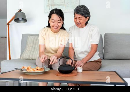 Una madre e una figlia cinesi condividono un momento caloroso nel loro salotto, sedute a un tavolino con tè e snack, incarnando l'amore familiare e la coesione Foto Stock