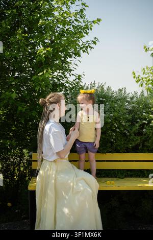 Una madre e sua figlia condividono un momento tenero in una giornata estiva soleggiata in un parco verdeggiante. Si siedono su una panchina gialla, circondate da fiori in fiore an Foto Stock