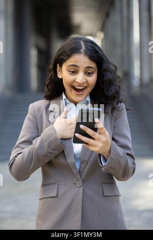 Donna professionista che reagisce con entusiasmo e sorpresa mentre guarda lo schermo del suo smartphone, ricevendo notizie positive o un messaggio di successo in un ambiente urbano all'aperto Foto Stock