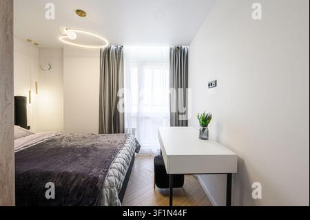 camera da letto moderna con un letto con una morbida coperta viola, una scrivania bianca e un'illuminazione elegante Foto Stock