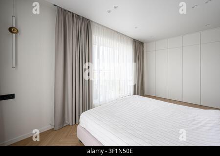 camera da letto minimalista con un letto con biancheria da letto bianca, lunghe tende grigie, un grande armadio bianco, pareti chiare e pavimenti in legno Foto Stock