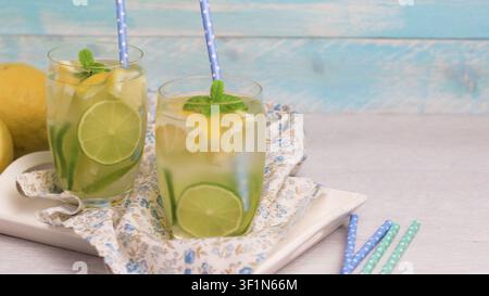 Due bicchieri trasparenti pieni di limonata fatta in casa, ghiaccio, fette di lime fresche e rametti di menta sono appoggiati su un vassoio bianco foderato con un panno floreale blu contro un Foto Stock