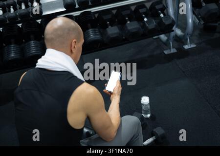Atleta che utilizza uno smartphone mentre riposa accanto ai manubri in palestra Foto Stock