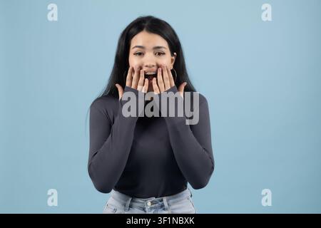 Giovane donna sorpresa che guarda la telecamera con le mani sul viso in studio Foto Stock