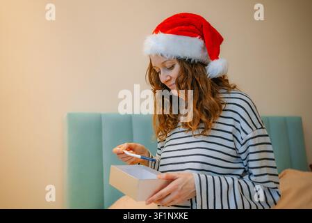 Donna con un cappello festivo di Babbo Natale che prepara un regalo di Natale premuroso, rivela un risultato positivo del test di gravidanza come una gioiosa sorpresa e annuncia al marito la futura genitorialità Foto Stock