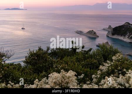 Paesaggio marino calmo su una spiaggia greca al crepuscolo con formazioni rocciose uniche, una barca a vela in navigazione e vegetazione costiera lussureggiante Foto Stock