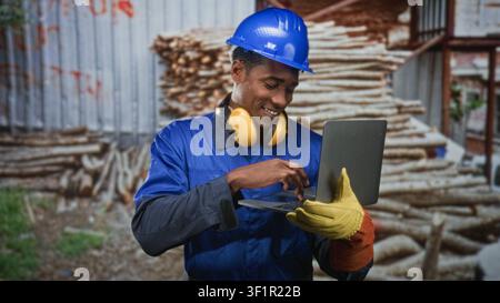 Operaio edile con casco protettivo tiene un laptop e digita con la mano guantata mentre sorride in un edificio; fiducia e produttività. Foto Stock