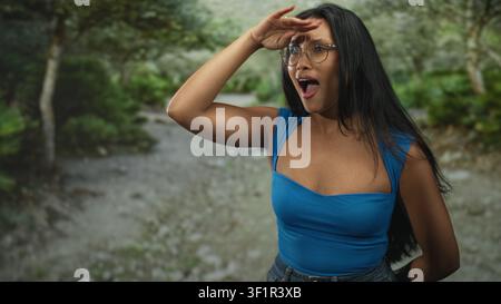 Donna che protegge gli occhi con la mano e punta a terra, indossa la cima blu e gli occhiali su un sentiero nella foresta; scoperta a sorpresa. Foto Stock
