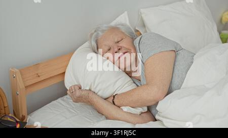 Una donna anziana abbraccia un cuscino mentre dorme su un letto in una camera da letto, distesa sul fianco con gli occhi chiusi e le mani che stringono il cuscino; comfort serenità. Foto Stock