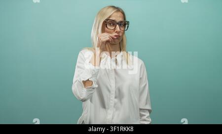 Donna bionda di mezza età con un dito sulle labbra per il silenzio in studio, indossa occhiali oversize e una camicia bianca su uno sfondo verde acqua; contenimento giocoso. Foto Stock