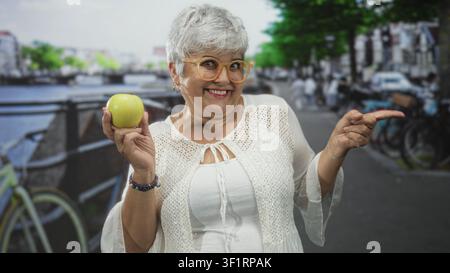 Donna anziana con capelli grigi e occhiali che tiene la mela verde in mano punta il dito su una strada della città sorridente; benessere gioioso. Foto Stock