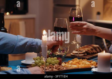 Coppia giovane che brinda con bicchieri di vino rosso a cena a un tavolo in cucina, con cena romantica, bicchieri di vino e tavolo da pranzo durante una cena calda in coppia o un momento di anniversario. Foto Stock