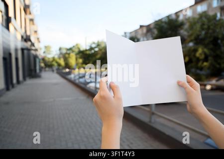 Mani che tengono una brochure o volantino bianco piegato vuota come modello all'aperto sul marciapiede di una strada cittadina Foto Stock