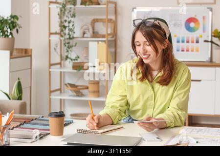 Giovane arredatrice sorridente che lavora a un progetto alla sua scrivania con campioni di tessuti e una tavolozza di colori, tiene campioni di tessuti e prende appunti Foto Stock