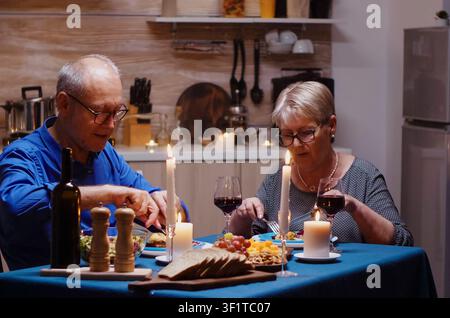 Coppia di anziani seduta a un tavolo da cucina accogliente con bicchieri di vino rosso, godendosi una cena allegra di anniversario insieme a casa, con vino, cibo e dettagli del tavolo che creano un'atmosfera calda di anniversario. Foto Stock