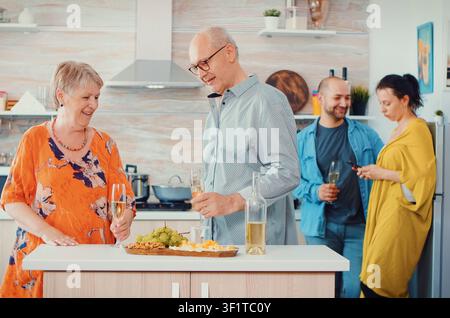 Adulti anziani allegri si abbracciano, ridono e ballano con bicchieri di vino bianco in cucina mentre una giovane coppia si trova nelle vicinanze, mentre cibo e dettagli domestici mantengono il romanticismo radicato nella vita domestica. Foto Stock