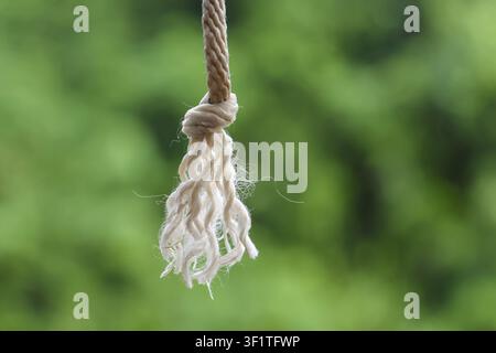Una nappina di corda di cotone sfilacciata appesa a un cordone annodato con morbide fibre naturali e sfondo sfocato di fogliame verde, texture rustica pacifica e primo piano Foto Stock