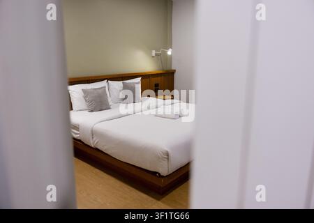 Una camera d'albergo con letto matrimoniale ordinatamente rifatto, lenzuola bianche, due cuscini grigi, lampada da comodino e asciugamani piegati che creano un'atmosfera calma e accogliente Foto Stock