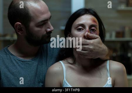 Moglie maltrattata affronta un partner aggressivo a casa mentre la copertura della bocca e la paura mostrano molestie, lesioni e rischi per la sicurezza personale. Foto Stock