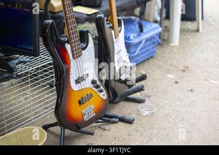 Un basso elettrico Sunburst e una chitarra elettrica nera su supporti in un mercato all'aperto con attrezzatura musicale e cassa, scena casual con toni caldi e rela Foto Stock