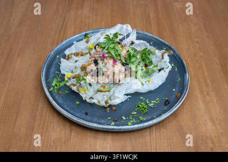 Tartara gourmet su una grande crosta bianca e croccante a forma di pizzo, guarnita con caviale nero, uova di pesce arancione, rucola e fiori commestibili su un piatto blu scuro. Foto Stock