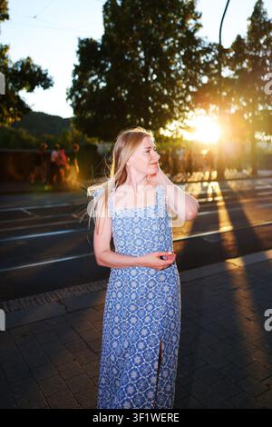 Bella giovane donna in un vestito estivo blu che cammina all'ora dorata in una città europea, sorridendo sopra la spalla nella calda luce del tramonto. Foto Stock