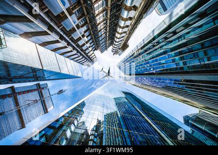Vista ad angolazione bassa che guarda direttamente verso la skyline del distretto finanziario nel centro di Londra con un aereo nel cielo che passa Foto Stock