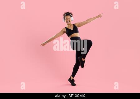 Donna nera atletica con cuffie che bilancia su una gamba e indica verso l'esterno Foto Stock