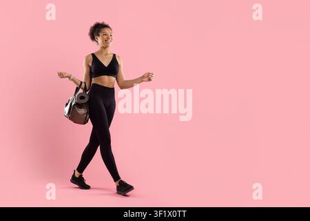 Donna nera in abbigliamento sportivo che cammina con una borsa da palestra e un tappeto da yoga arrotolato Foto Stock