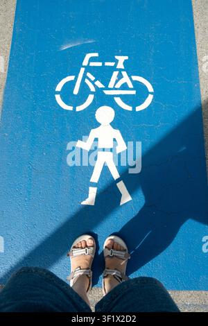 Vista POV dall'alto delle gambe di una persona in piedi su una pista pedonale e ciclabile blu condivisa Foto Stock