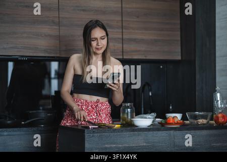 Giovane donna prepara un'insalata fresca nella sua cucina di casa seguendo una ricetta sul suo smartphone. Cucina sana, apprendimento culinario, cibo fatto in casa, Foto Stock