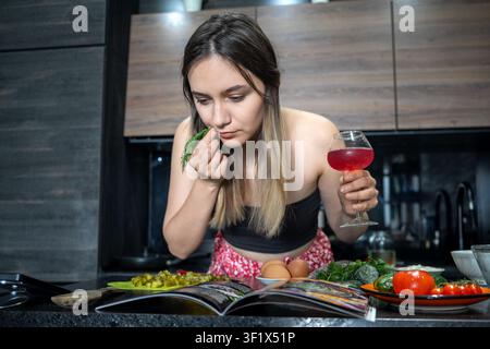 Giovane donna attraente prepara un'insalata fatta in casa in una cucina moderna seguendo una ricetta di libro di cucina. Cuoca principiante annusa l'aneto fresco e si gode uno spillo Foto Stock