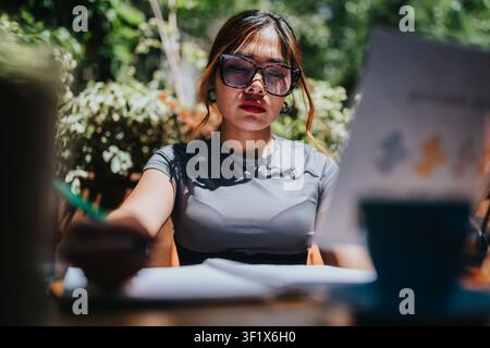 Giovane donna che studia all'aperto in un caffè, concentrata sulla lettura e sulla scrittura con gli occhiali Foto Stock