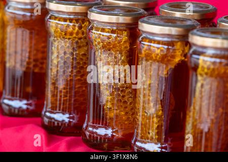 Primo piano di barattoli di vetro con miele selvatico crudo e trama visibile dei favi, liquido ambrato dorato su sfondo rosso Foto Stock