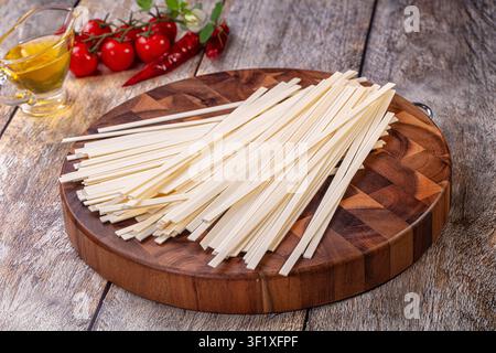 Maccaroni udon crudi disposti su un tagliere di legno rustico. Lo sfondo mostra pomodorini, peperoncini rossi, olio d'oliva in una brocca di vetro Foto Stock