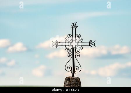 Una croce di ferro si erge su una pietra, sullo sfondo di un cielo blu chiaro con soffici nuvole bianche in un campo. Foto Stock