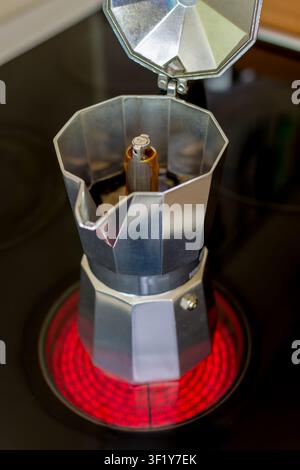 Moka in alluminio con coperchio aperto che prepara caffè scuro su un piano cottura elettrico rosso acceso in una cucina. Foto Stock