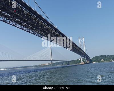 L'iconico Forth Road Bridge e il moderno Queensferry Crossing affiancati, catturati da una barca in una giornata estiva soleggiata in Scozia. Foto Stock