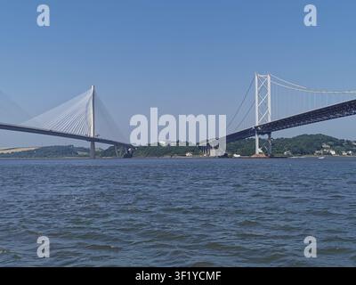 L'iconico Forth Road Bridge e il moderno Queensferry Crossing affiancati, catturati da una barca in una giornata estiva soleggiata in Scozia. Foto Stock