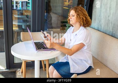 Donna di mezza età che lavora su laptop e telefono in un caffè, con aria concentrata e pensierosa Foto Stock